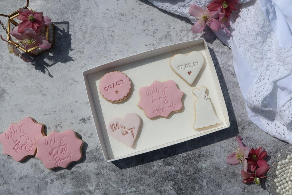 Will you be my bridesmaid proposal cookie set, pinks and rose gold, wedding dress cookie, heart cookie with wedding date, heart with bride and groom initials, round cookie with recipients name. Will you be my flower girl cookie and will you be my maid of honour cookie to side